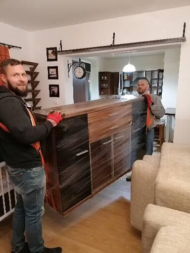 Dwóch mężczyzn w rękawicach ochronnych i pasach transportowych przenosi owiniętą folią komodę w jasnym wnętrzu mieszkania, widoczny zegar dworcowy na ścianie.