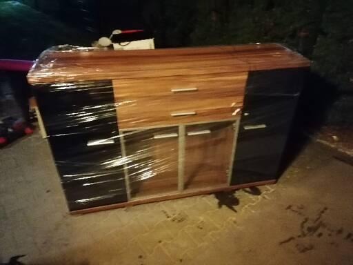 Komoda z drewnianymi frontami i czarnymi bokami, owinięta folią stretch, przygotowana do transportu. Widoczne ślady użytkowania na podłożu.