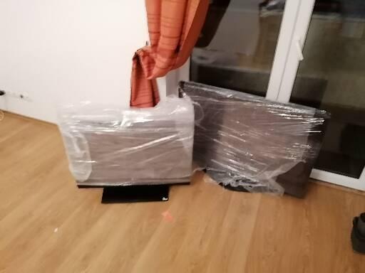 Dwa telewizory owinięte folią stretch, przygotowane do transportu podczas przeprowadzki, ustawione na drewnianej podłodze w pokoju z pomarańczową zasłoną i białym oknem.