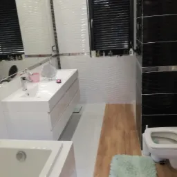 Nowoczesna łazienka z białymi, falistymi płytkami, czarnymi akcentami i drewnopodobną podłogą; widoczna umywalka z szafką, wanna, toaleta i okno z żaluzjami.