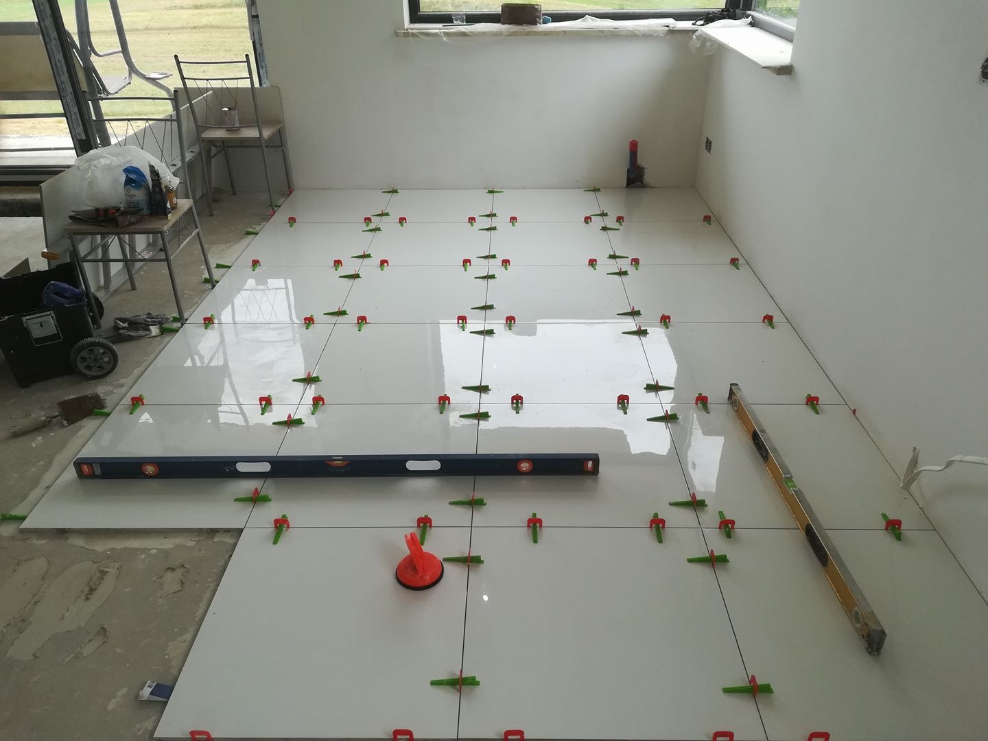 Białe, błyszczące płytki podłogowe w trakcie układania, z użyciem poziomicy i systemu poziomowania płytek (czerwone i zielone kliny), widoczne narzędzia pracy i częściowo przygotowane podłoże.