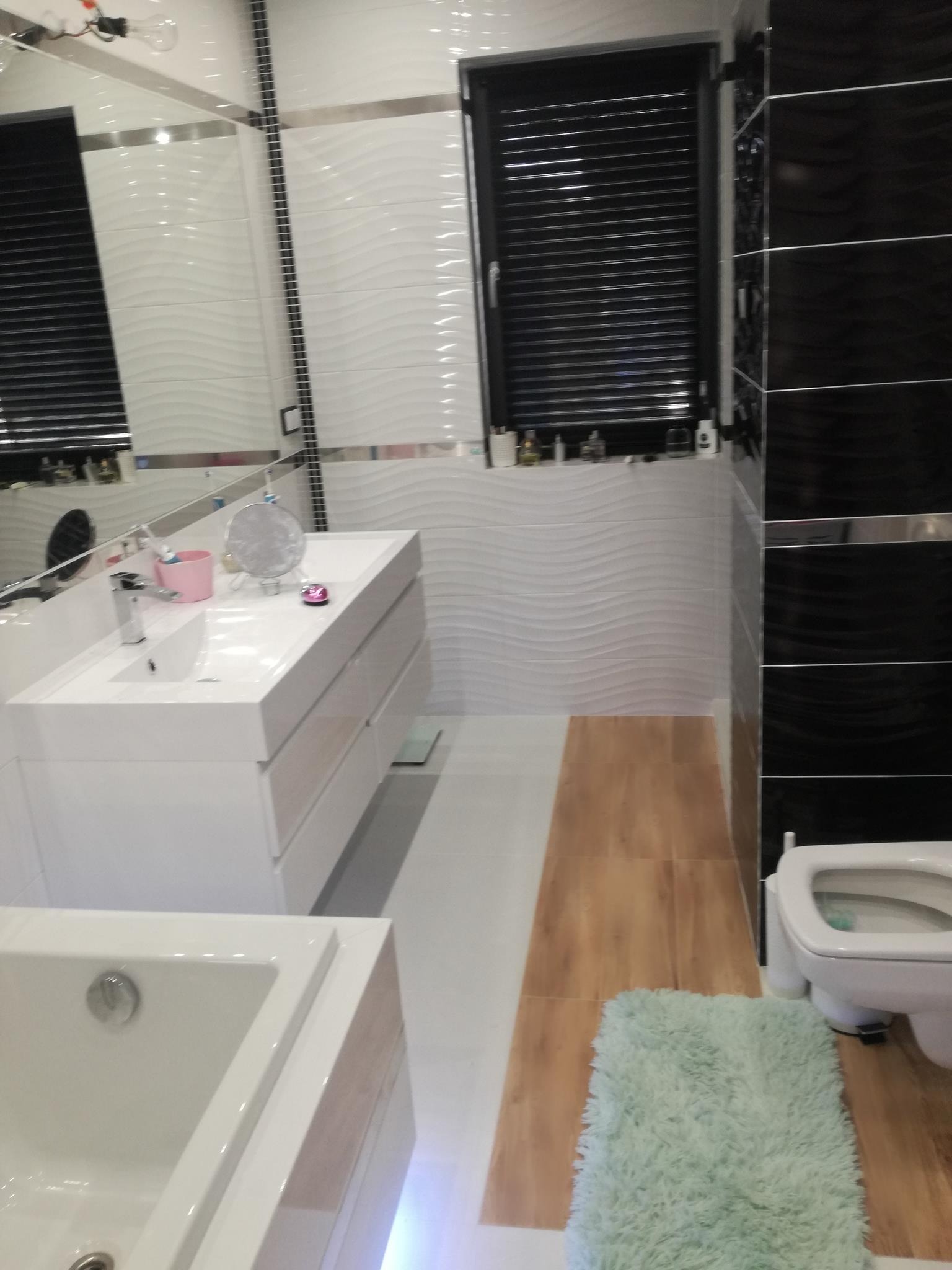 Nowoczesna łazienka z białymi, falistymi płytkami, czarnymi akcentami i drewnopodobną podłogą; widoczna umywalka z szafką, wanna, toaleta i okno z żaluzjami.