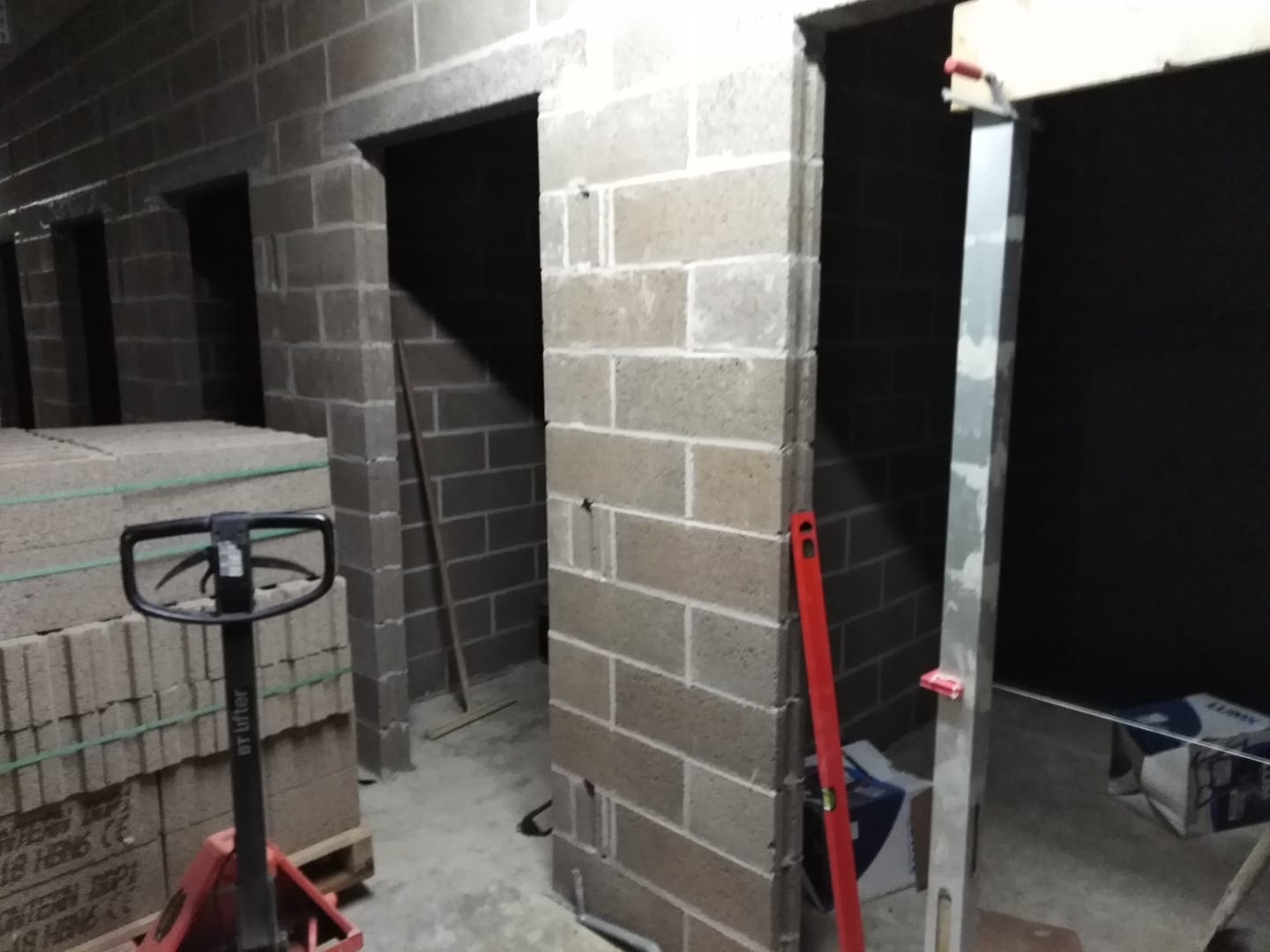 Wznoszenie ścian działowych z bloczków betonowych, widoczne surowe ściany z otworami drzwiowymi, paleta bloczków i wózek paletowy.