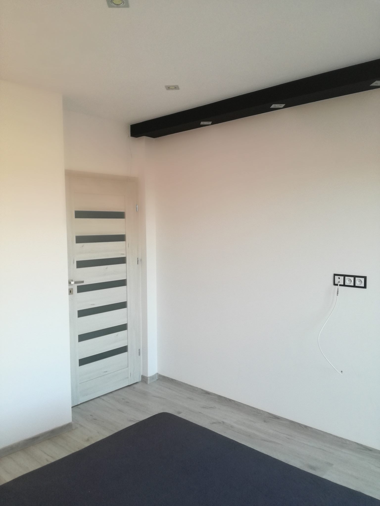 Wnętrze pokoju po remoncie: świeżo pomalowane białe ściany, jasna podłoga imitująca drewno, nowoczesne gniazdka elektryczne z podłączonym przewodem, białe drzwi z szarymi poziomymi paskami, czarna...