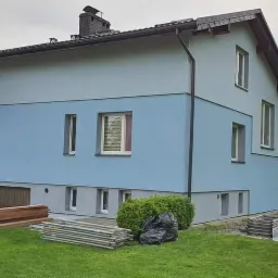 Dwukondygnacyjny dom z elewacją w dwóch odcieniach niebieskiego, z widocznymi rusztowaniami, stertą desek, workami na śmieci i zaparkowanym busem, sugerujący prace wykończeniowe.