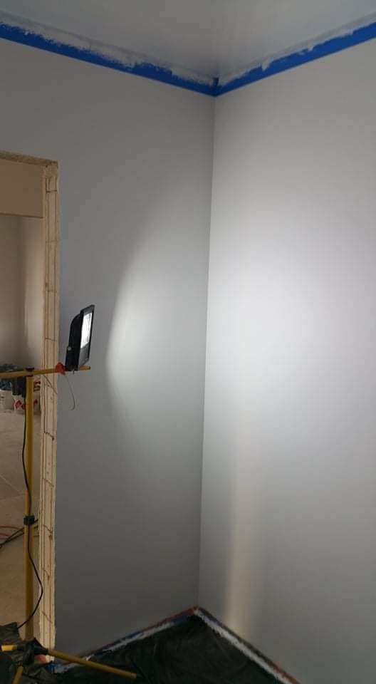 Świeżo pomalowany róg pokoju z niebieską taśmą malarską przy suficie, lampa halogenowa oświetla ścianę, podłoga zabezpieczona folią malarską.