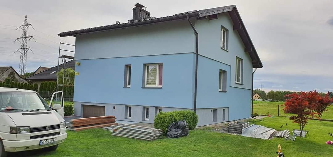 Dwukondygnacyjny dom z elewacją w dwóch odcieniach niebieskiego, z widocznymi rusztowaniami, stertą desek, workami na śmieci i zaparkowanym busem, sugerujący prace wykończeniowe.