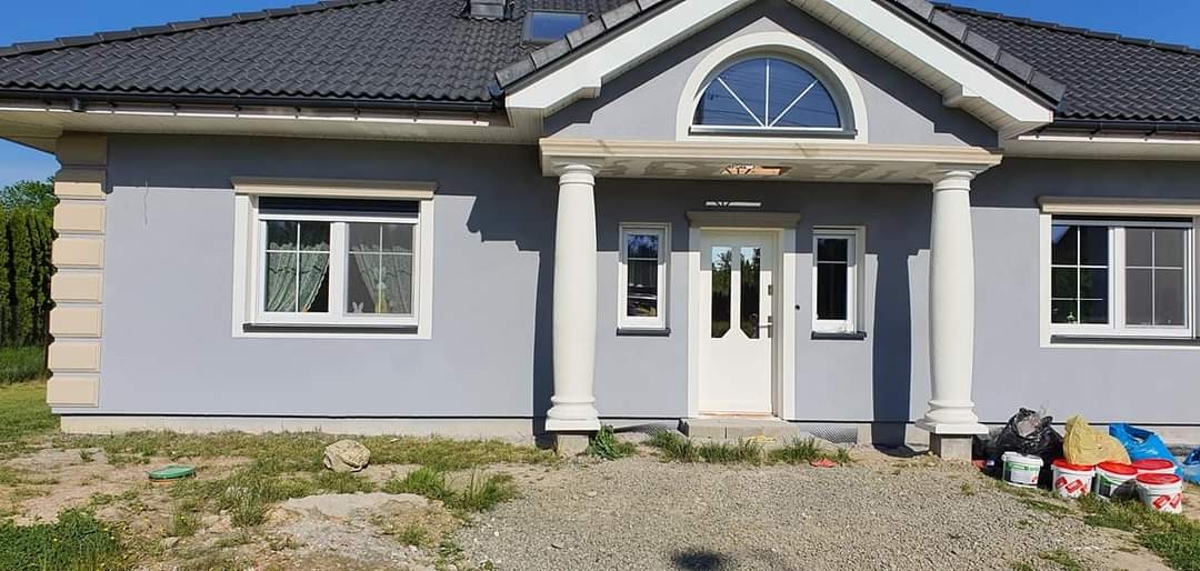 Dom jednorodzinny z szarą elewacją, białymi kolumnami i oknami, widoczne puszki po farbie i worki na podłożu.