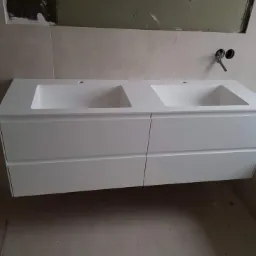 Biała szafka łazienkowa podwieszana z dwoma umywalkami, widoczne niedokończone prace wykończeniowe na ścianie i brak baterii.