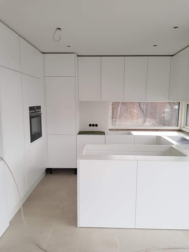 Montaż białej kuchni na wymiar z wyspą kuchenną i wbudowanym piekarnikiem, widoczne niedokończone elementy i okno w tle.