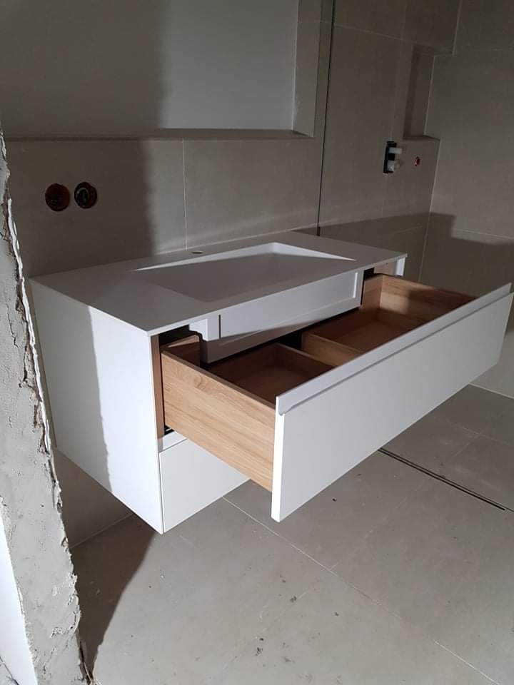 Biała szafka łazienkowa pod umywalkę z wysuniętą szufladą z jasnego drewna, widoczne niedokończone wykończenie ściany, szare płytki podłogowe.