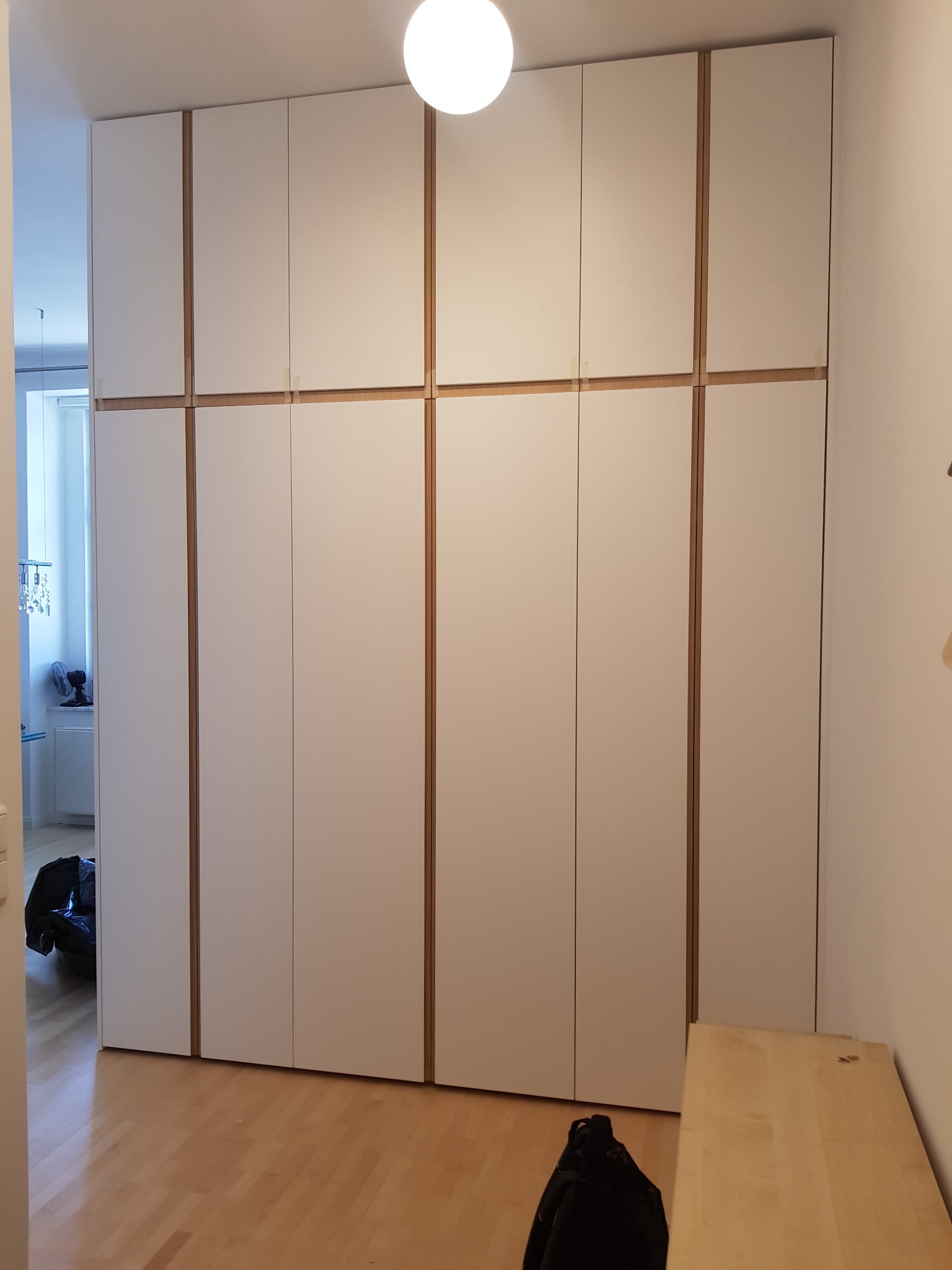Duża, biała szafa wnękowa z drewnianymi akcentami, zajmująca całą ścianę, widoczna z perspektywy podłogi, z fragmentem drewnianego blatu po prawej stronie i torbą na podłodze.