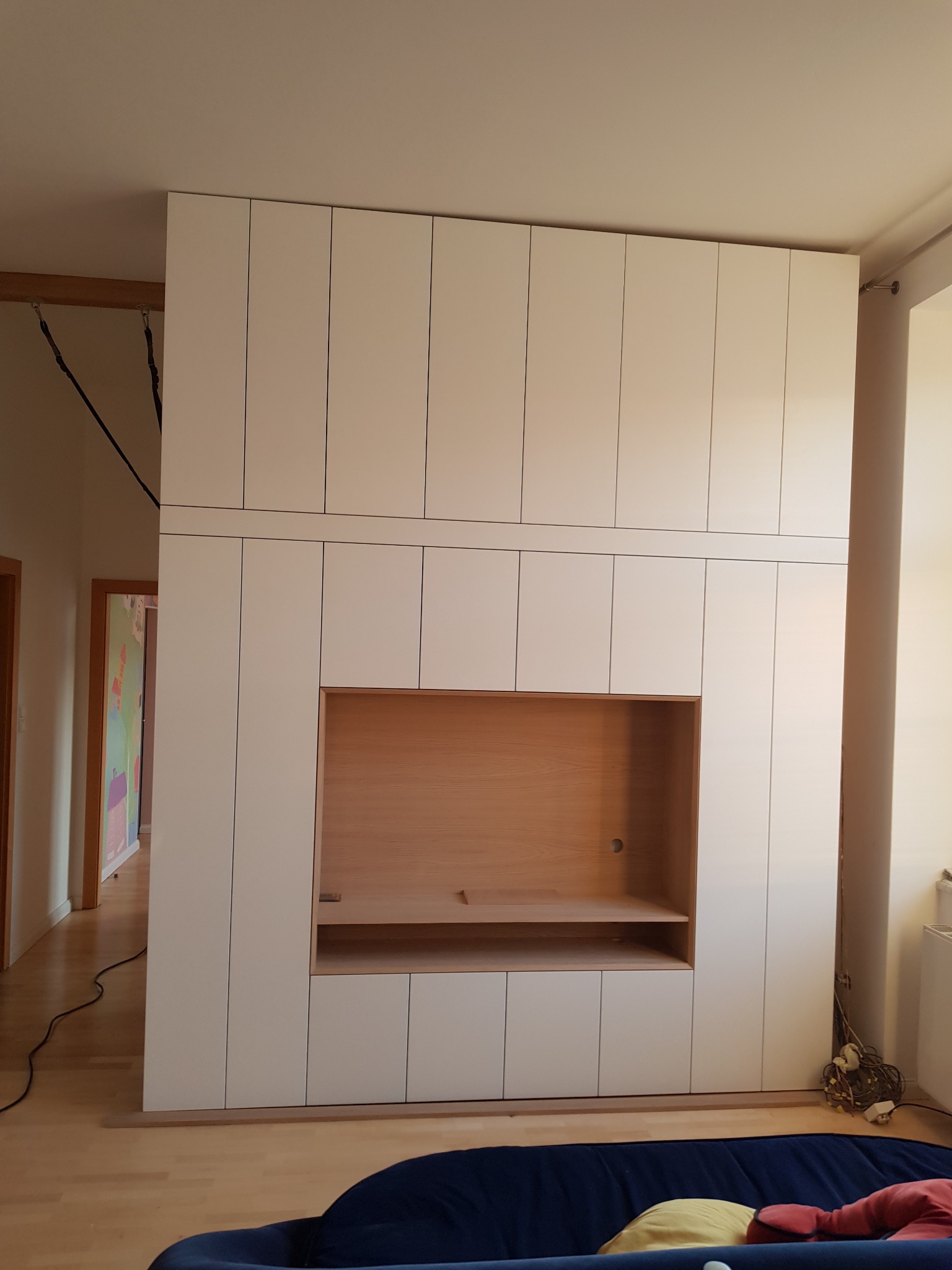 Biała zabudowa ściany z szafkami i wnęką na telewizor, wykończoną jasnym drewnem, widoczna z boku w salonie.