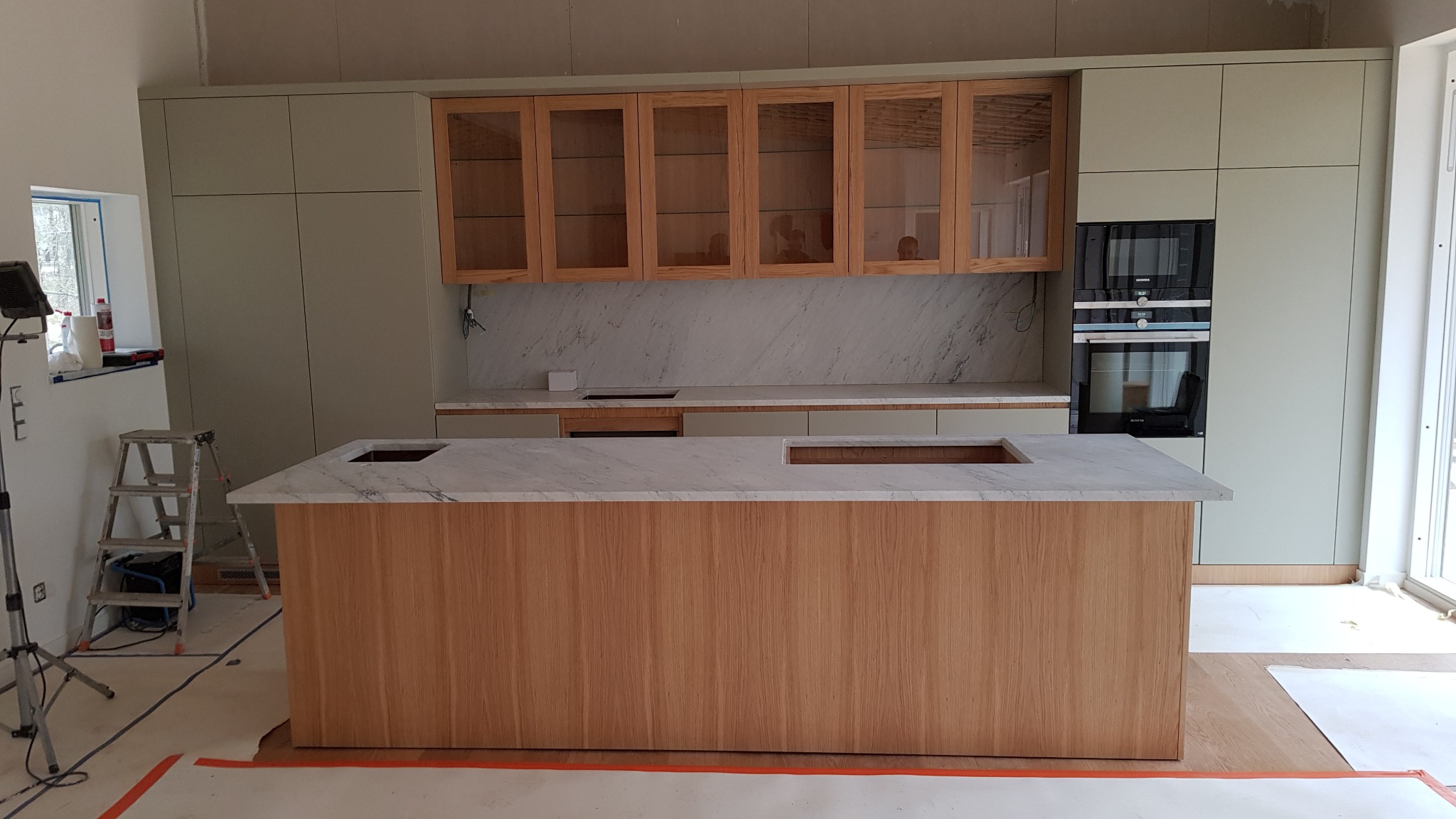 Wnętrze kuchni w trakcie montażu, widoczna wyspa z marmurowym blatem i drewnianym wykończeniem, szafki wiszące z przeszklonymi frontami, zabudowa w kolorze pistacjowym, sprzęt AGD w zabudowie...
