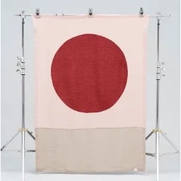 Dekoracyjna tkanina z dużym czerwonym kołem na różowym tle i beżowym pasem u dołu, zawieszona na metalowym statywie w studio.