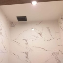 Bart-Bud - Białe płytki imitujące marmur na ścianie łazienki, z widoczną kratką wentylacyjną i oświetleniem sufitowym. Minimalistyczny design, jasne wnętrze.