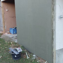 Bart-Bud - Ściana budynku po nałożeniu szarej warstwy tynku, z widocznymi narzędziami pracy i fragmentami ocieplenia na trawie. Widoczne siatka zbrojeniowa i styropian.