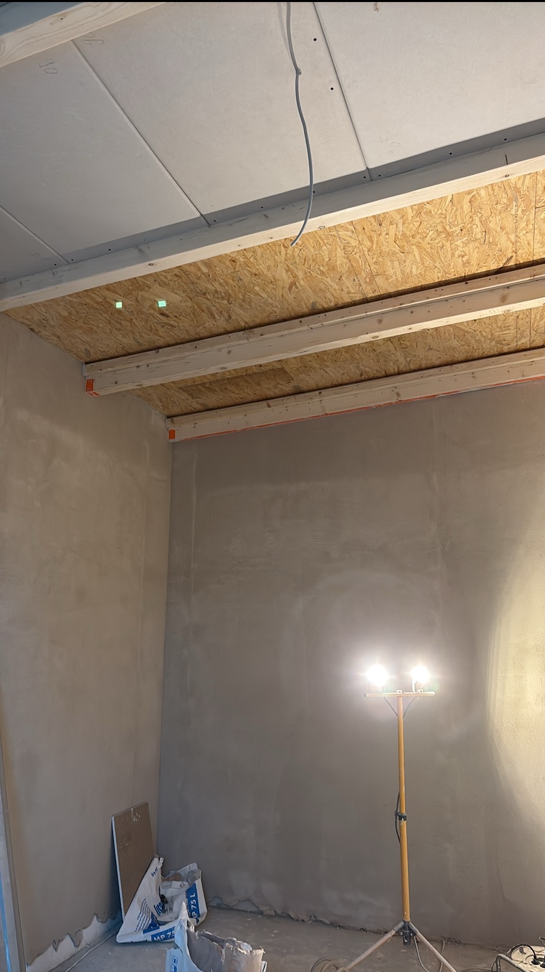 Wnętrze w trakcie prac wykończeniowych: surowe ściany po szpachlowaniu, widoczne płyty OSB na suficie i lampa budowlana oświetlająca pomieszczenie.