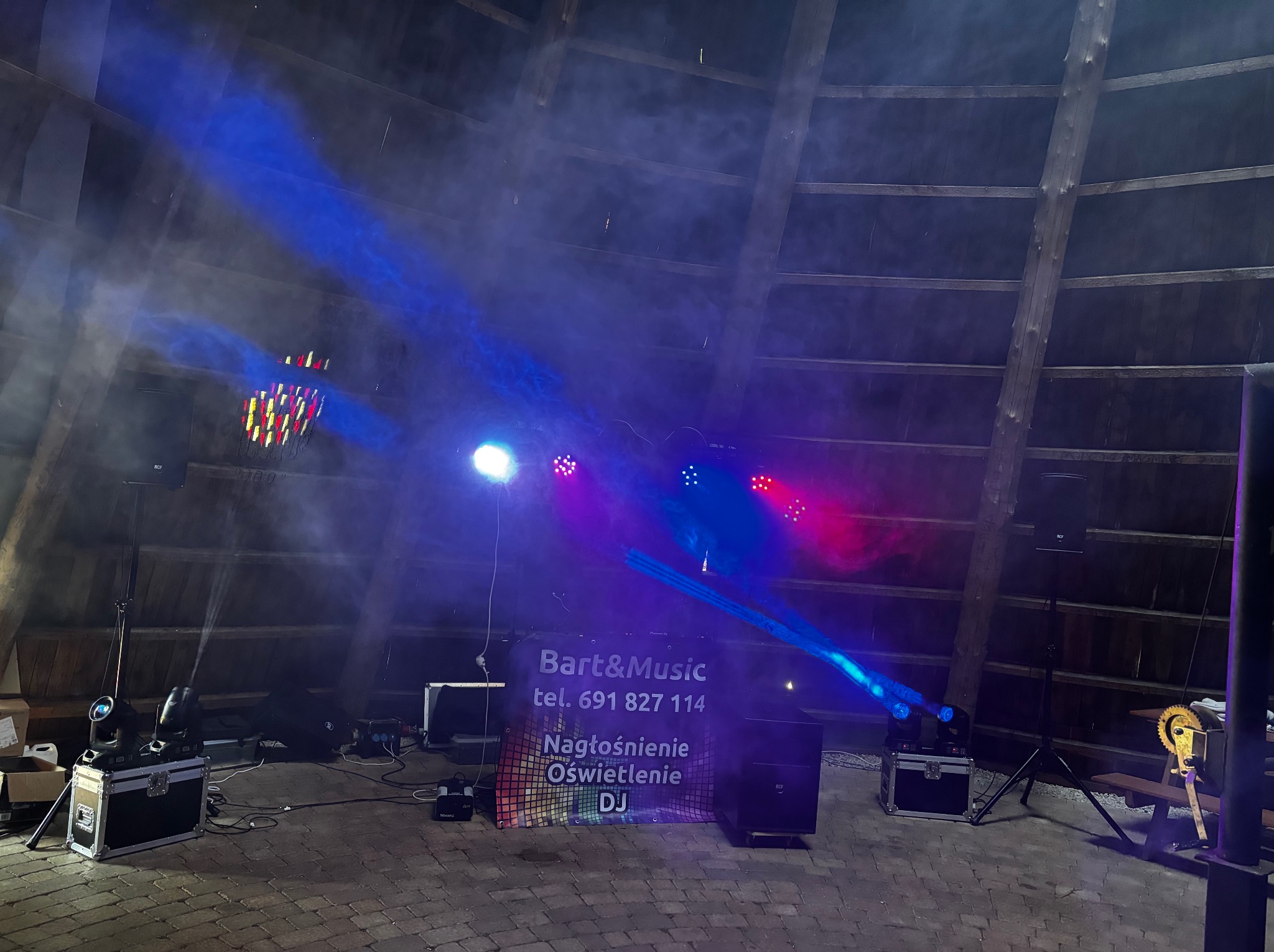 Scena z nagłośnieniem i oświetleniem DJ-a Bart&Music, z intensywnymi niebieskimi i czerwonymi światłami, w rustykalnym wnętrzu z drewnianą konstrukcją dachu.