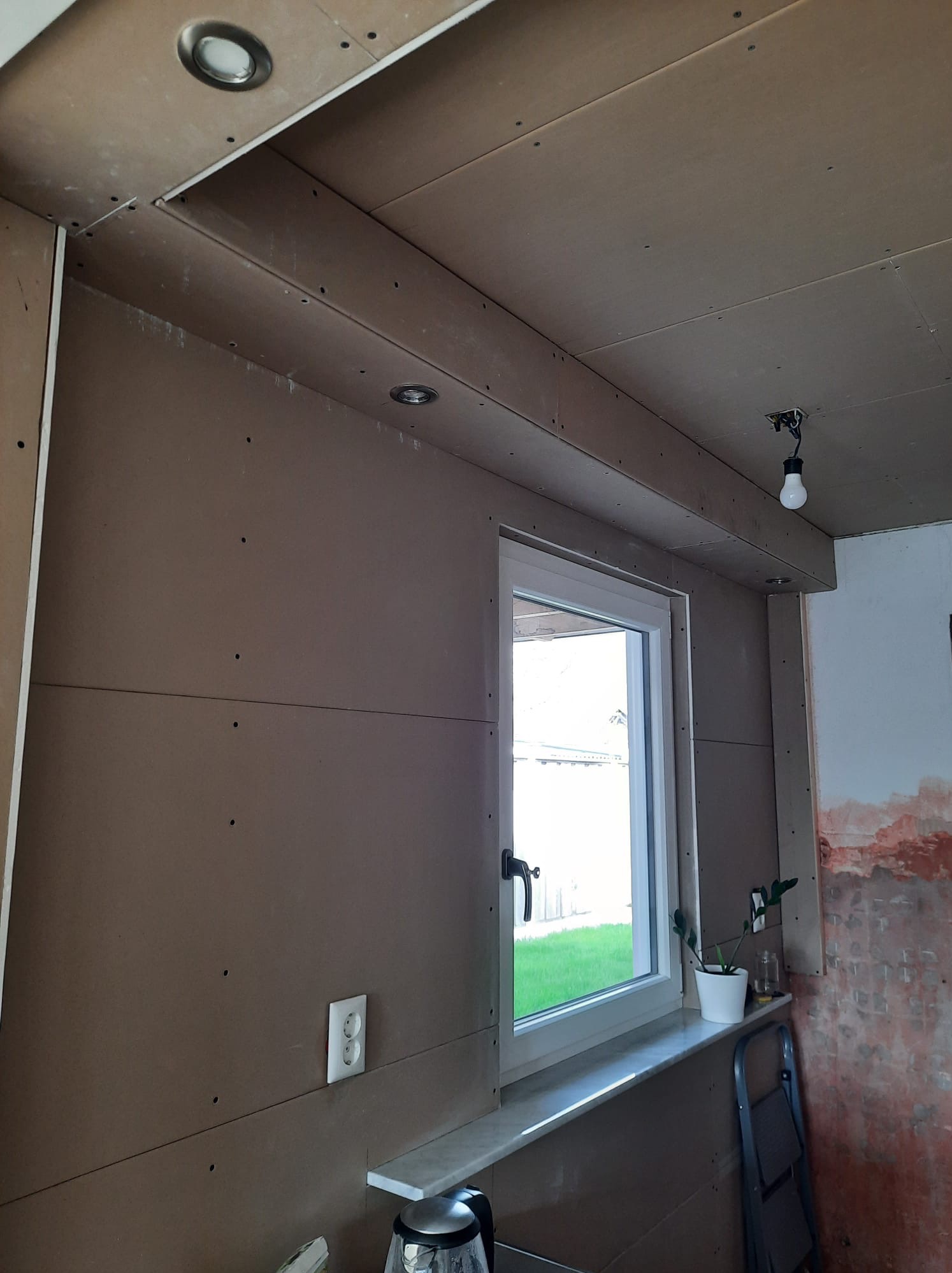 Wnętrze w trakcie prac: ściany i sufit pokryte płytami kartonowo-gipsowymi, widoczne punkty świetlne, okno z parapetem i fragment ściany z resztkami starej farby.