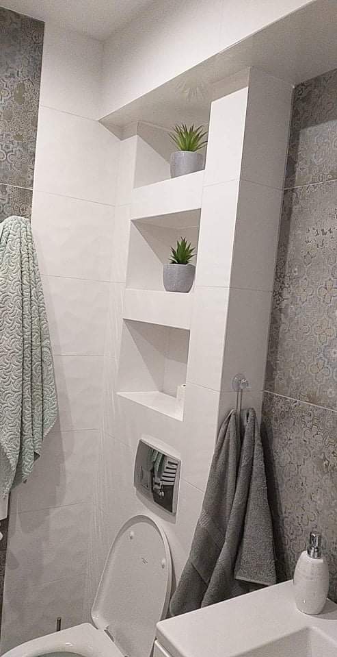 Nowoczesna łazienka z wnęką wykończoną płytkami, w której umieszczono dekoracyjne rośliny. Widoczne płytki w dwóch odcieniach szarości i wiszące ręczniki.