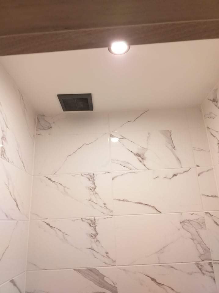 Białe płytki imitujące marmur na ścianie łazienki, z widoczną kratką wentylacyjną i oświetleniem sufitowym. Minimalistyczny design, jasne wnętrze.