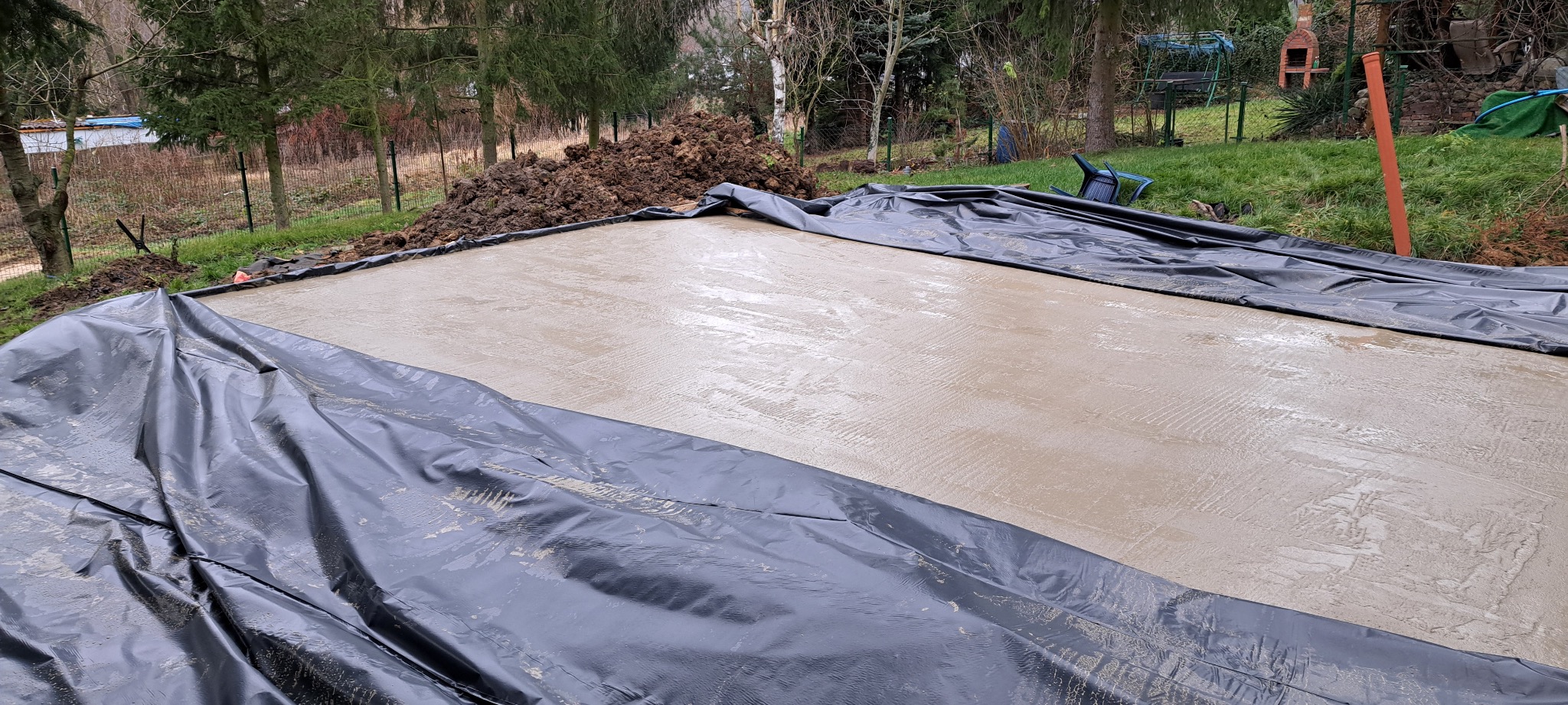 Wylana, świeża wylewka betonowa na zewnątrz, zabezpieczona czarną folią. W tle widoczna sterta ziemi i zieleń ogrodu. Widok z góry, lekko z ukosa.