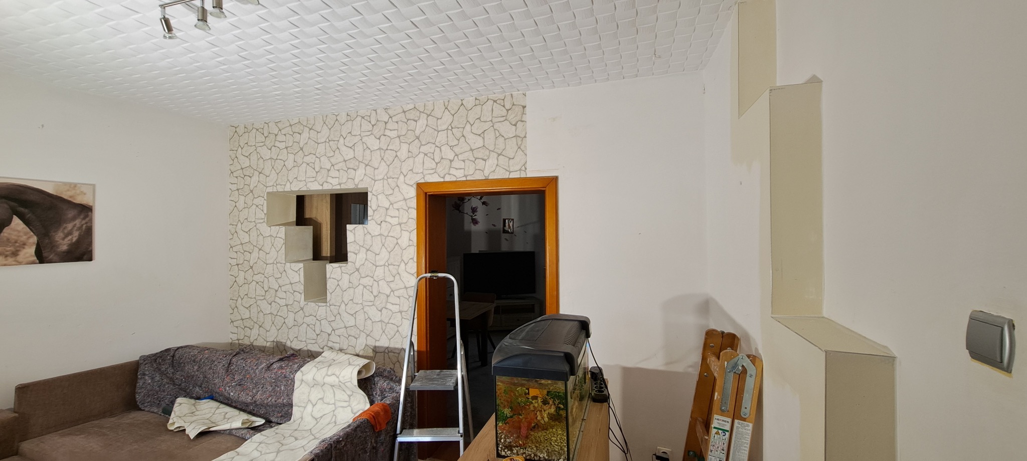 Pokój w trakcie wykańczania: ściana z tapetą imitującą kamień, otwór dekoracyjny, drabina, akwarium. Sufit z kasetonami. Widoczne narzędzia i elementy wyposażenia.