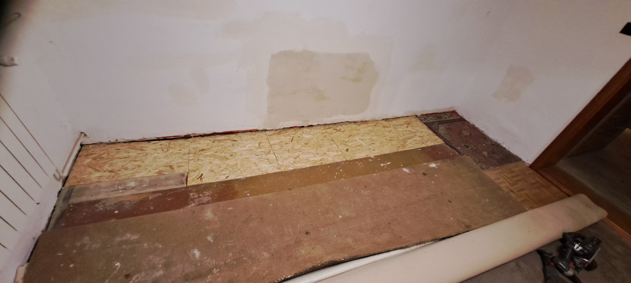 Fragment pokoju w trakcie remontu: ściana z łatami po szpachlowaniu, podłoga z fragmentami starej i nowej nawierzchni, zwinięty dywan. Widoczne narzędzia.