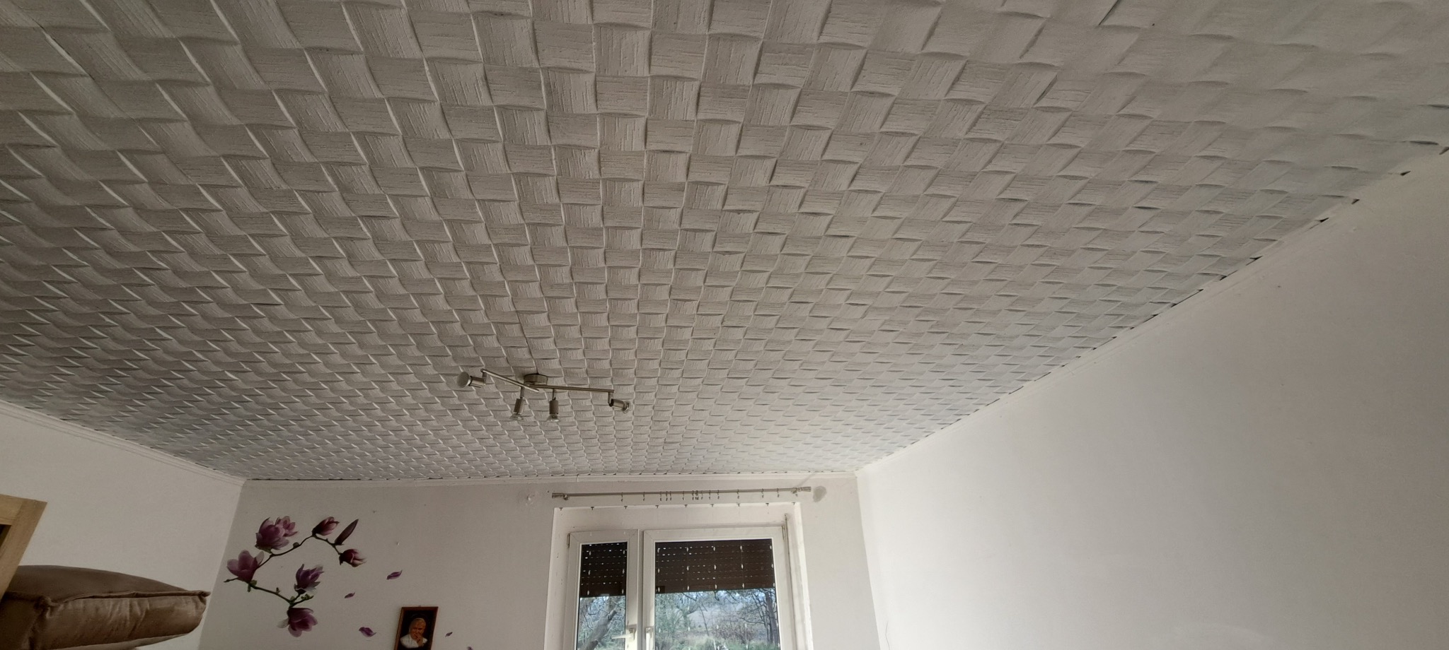 Dekoracyjny sufit z paneli PCV o wzorze plecionki, oświetlony lampą sufitową. Widoczne okno i fragment ściany z naklejką kwiatową.