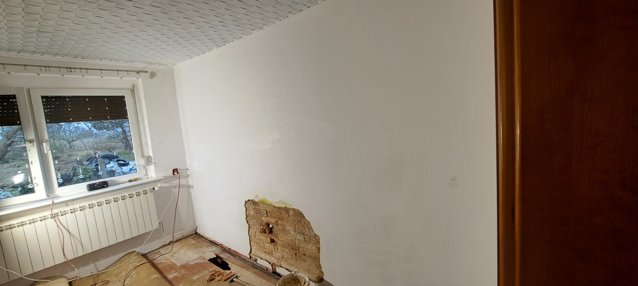 Pokój w trakcie remontu: ściana z odsłoniętą cegłą, częściowo zagruntowana, okno z roletami, grzejnik, podłoga z resztkami paneli, widoczny fragment drzwi.
