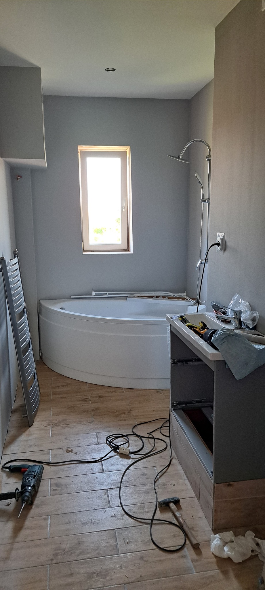 Wnętrze łazienki w trakcie remontu: narożna wanna, drewnopodobne płytki, częściowo zamontowana szafka pod umywalkę, narzędzia na podłodze. Widok na okno.