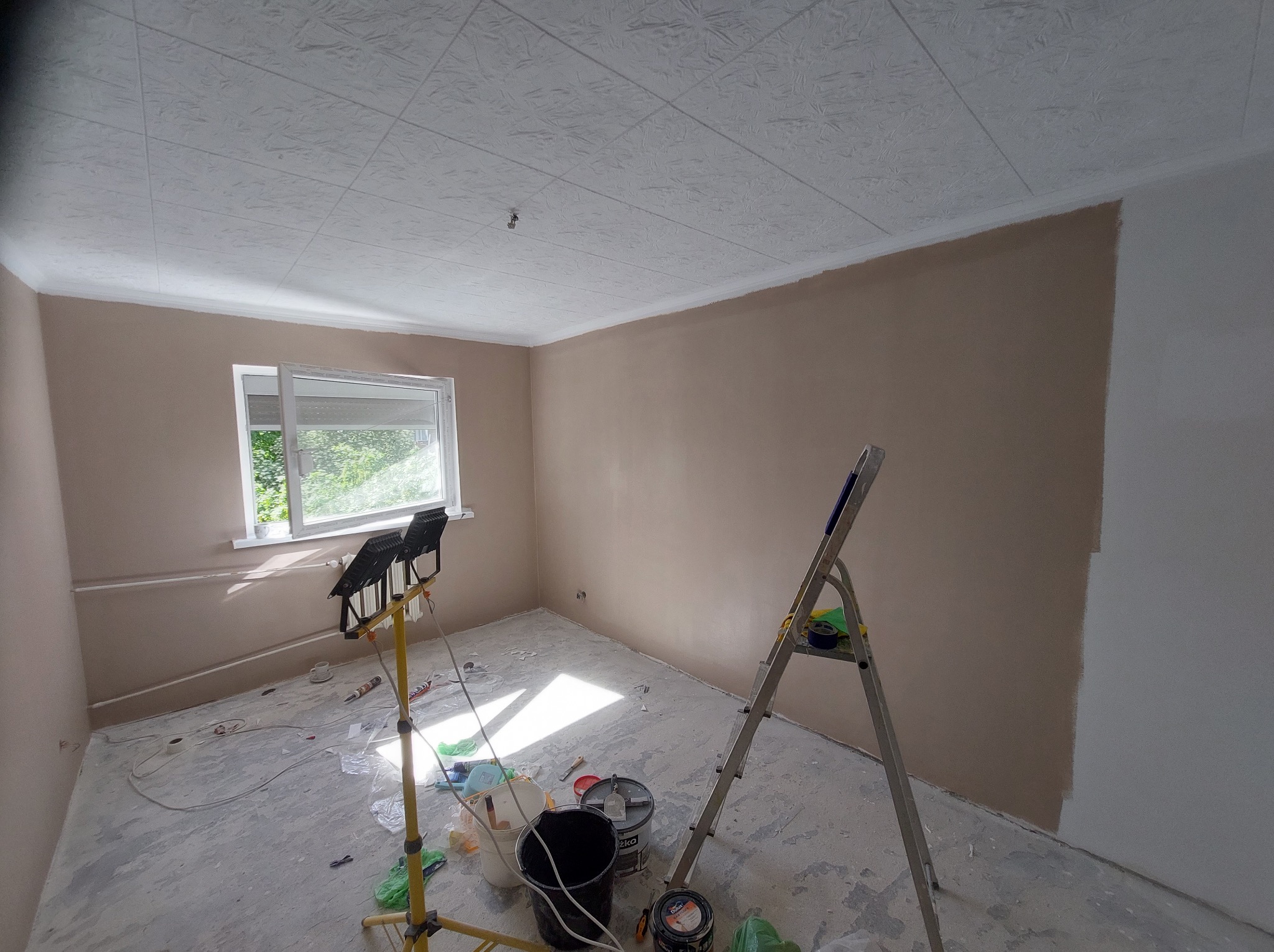 Pokój w trakcie malowania: ściany częściowo w kolorze beżowym, częściowo białe, drabina, lampa budowlana, wiadro z farbą. Sufit z fakturą. Widok na okno.