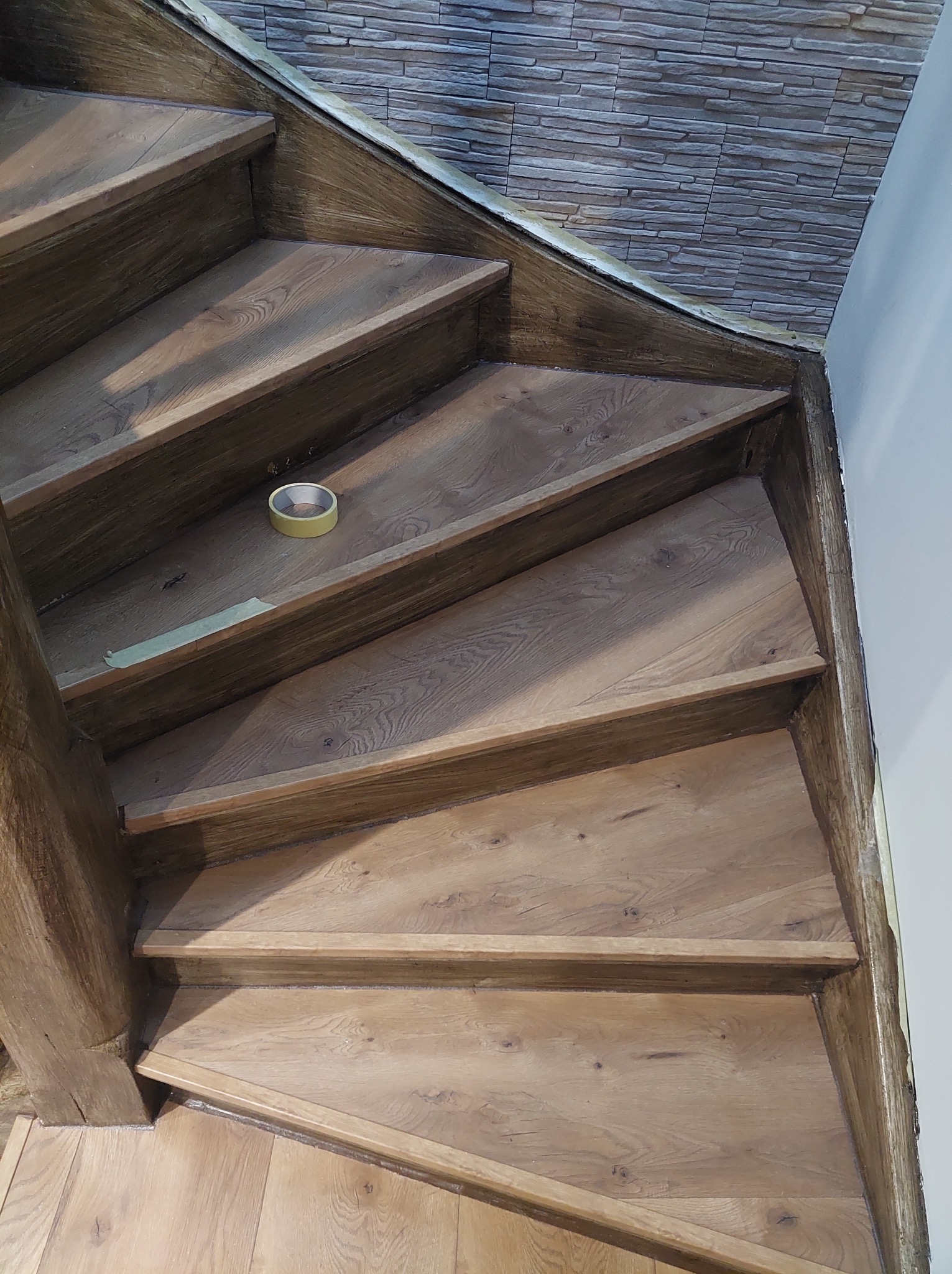 Drewniane schody w trakcie montażu, zabezpieczone taśmą malarską, z widoczną strukturą drewna i fragmentem ściany wykończonej imitacją kamienia.