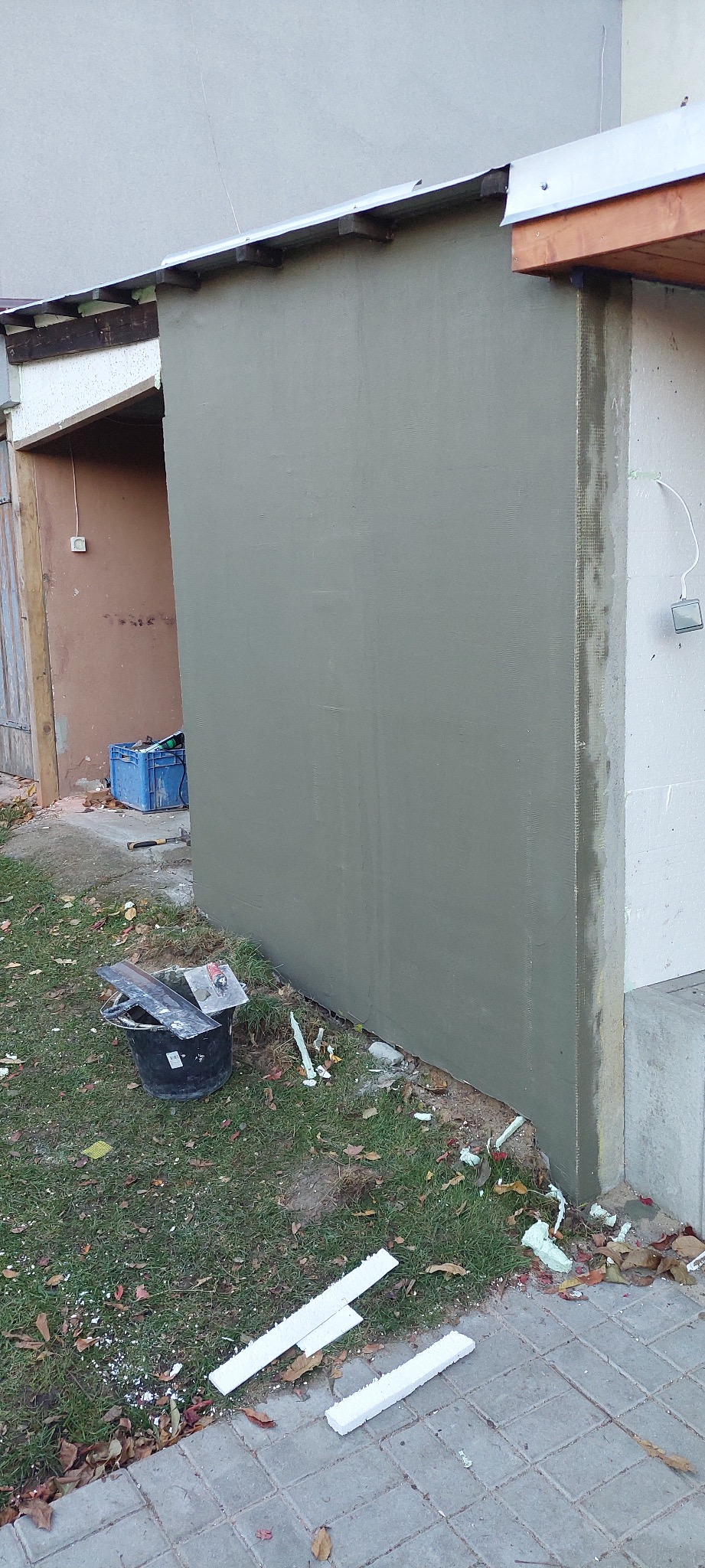 Ściana budynku po nałożeniu szarej warstwy tynku, z widocznymi narzędziami pracy i fragmentami ocieplenia na trawie. Widoczne siatka zbrojeniowa i styropian.