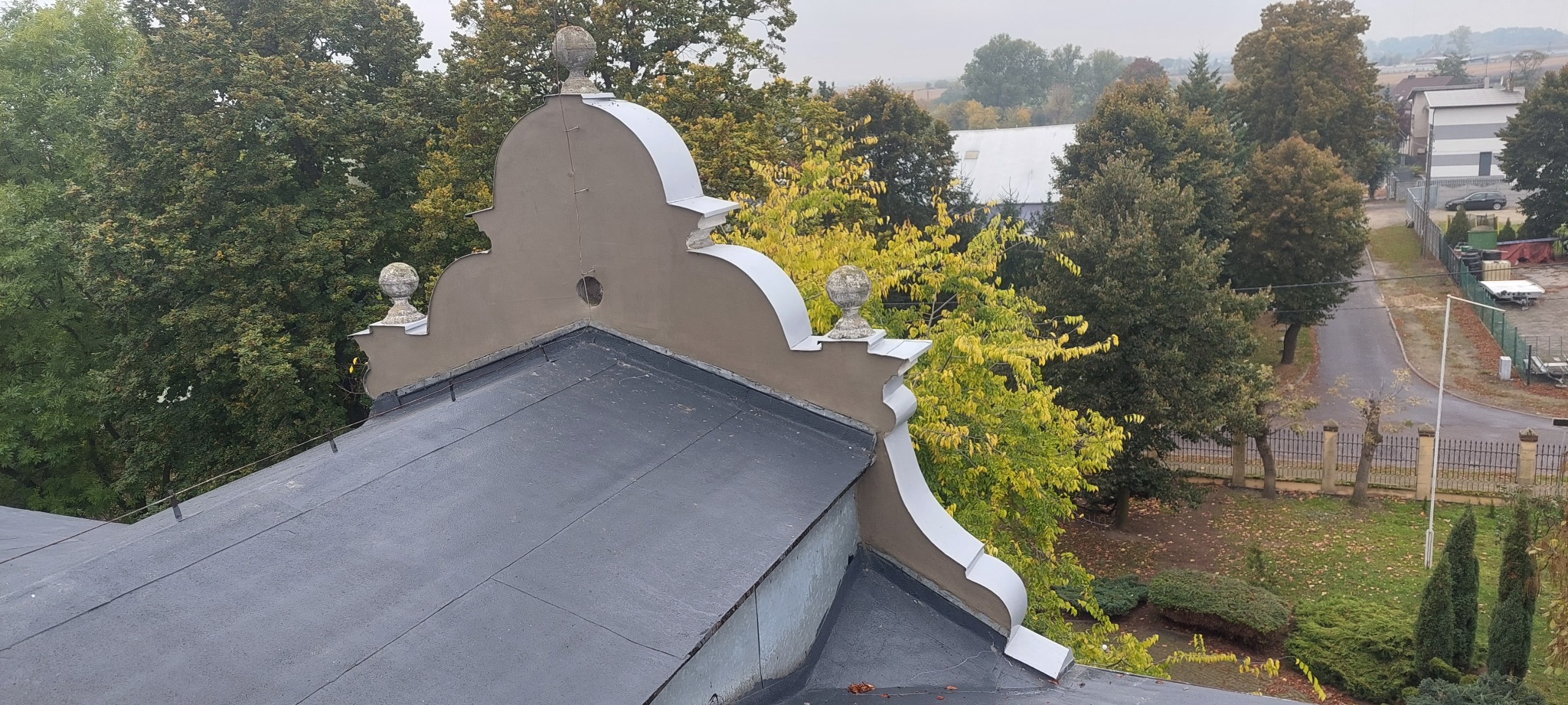 Szary dach z dekoracyjnym wykończeniem i kulami, na tle zieleni drzew i zabudowań. Widok z góry, pochmurny dzień.