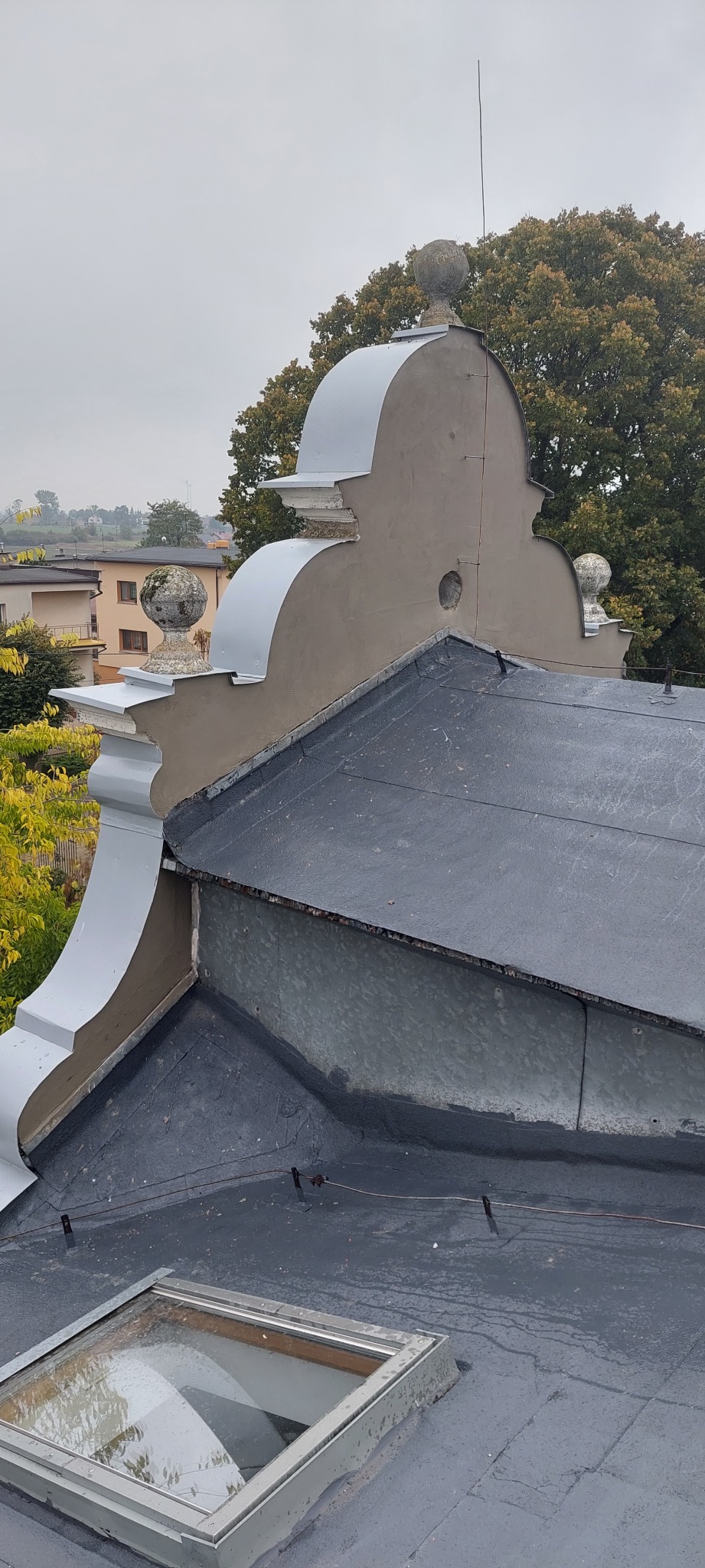 Szczegółowy widok na odnowioną, ciemną papę dachową z lukarną i ozdobnym wykończeniem w stylu dworkowym, zwieńczonym kulami. W tle zabudowa mieszkalna i zieleń.