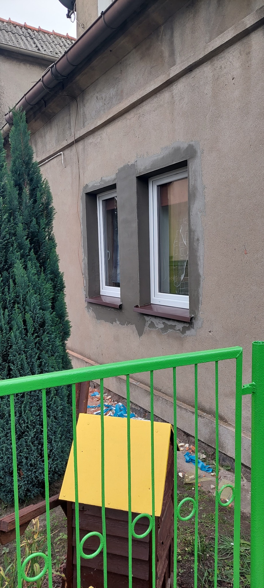 Dwa białe okna po wymianie, z widocznymi śladami świeżo nałożonej zaprawy na elewacji budynku. W tle zielony krzew i fragment zielonego ogrodzenia.