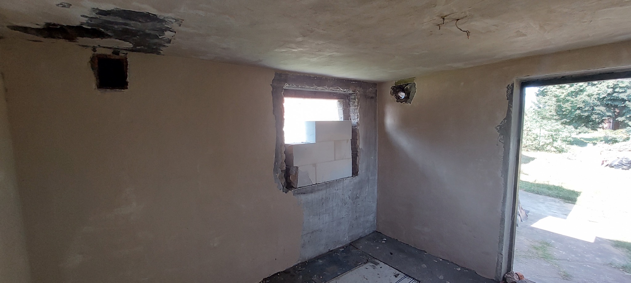 Surowe wnętrze w trakcie prac wykończeniowych: ściany po szpachlowaniu, częściowo zakryte okno z pustaków, widoczne instalacje w ścianach i niedoskonałości na suficie.