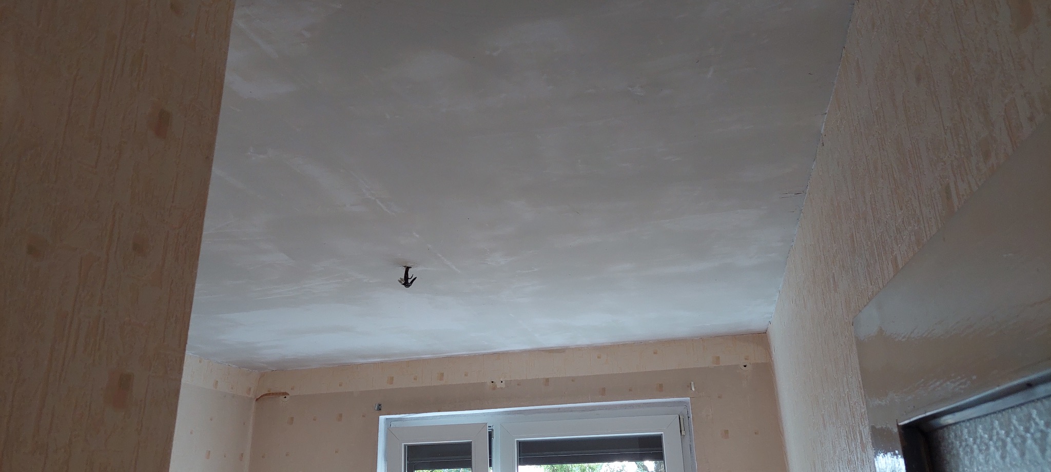 Wnętrze pokoju w trakcie remontu: gładzie na suficie, ściany z tapetą, okno. Widoczne ślady szpachlowania i przygotowania do malowania. Wystający kabel od oświetlenia.