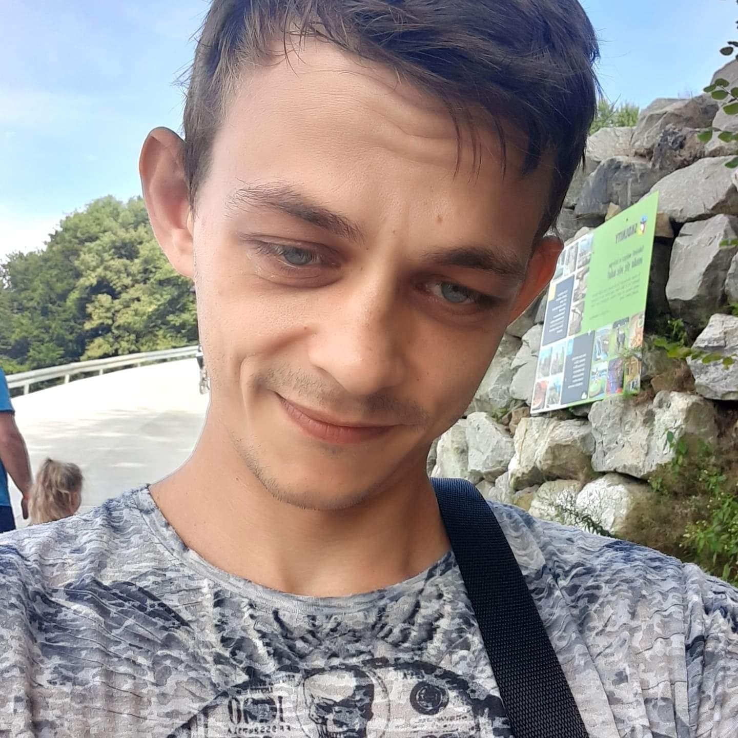 Młody mężczyzna z niebieskimi oczami, w koszulce z motywem czaszki, uśmiecha się do kamery na tle kamiennej ściany i tablicy informacyjnej.