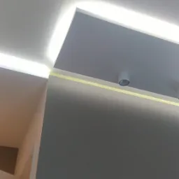 Nowoczesne oświetlenie sufitowe LED w narożniku pokoju, z widoczną taśmą malarską zabezpieczającą ścianę podczas prac wykończeniowych i pojedynczym punktowym reflektorem.