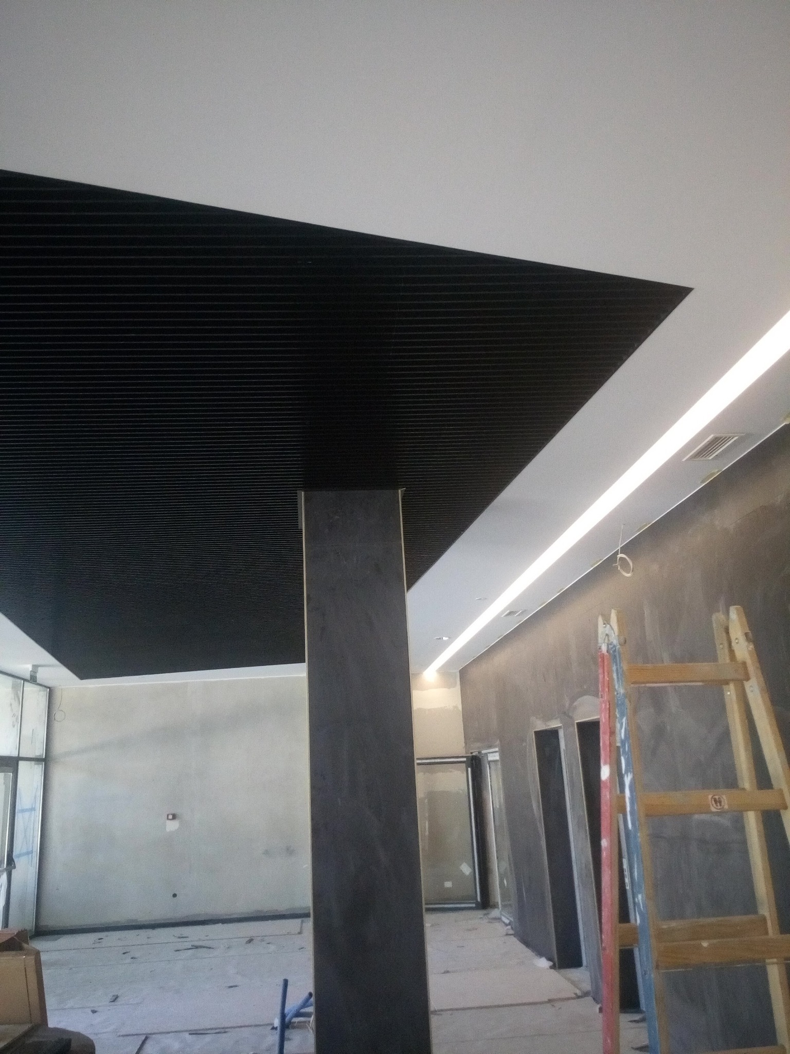 Częściowo zamontowany sufit rastrowy w biurze z widoczną kolumną, oświetleniem liniowym LED i drabiną w tle.