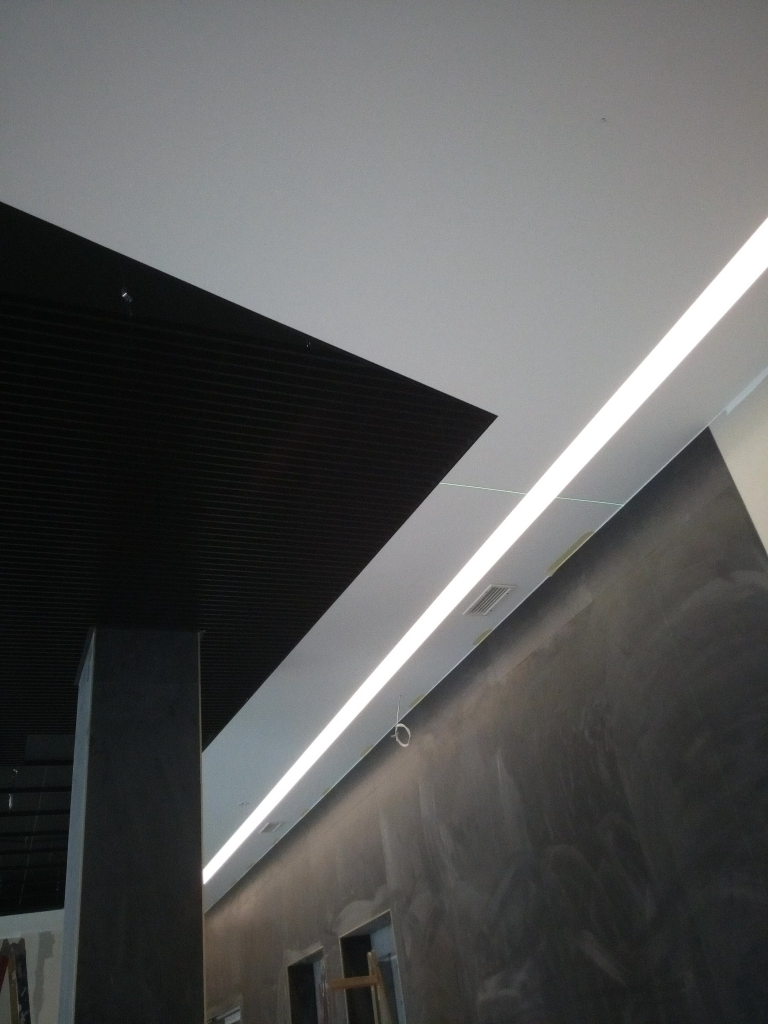 Nowoczesne wnętrze biurowe z czarnym, lamelowym sufitem podwieszanym i liniowym oświetleniem LED wzdłuż ściany o surowym wykończeniu.