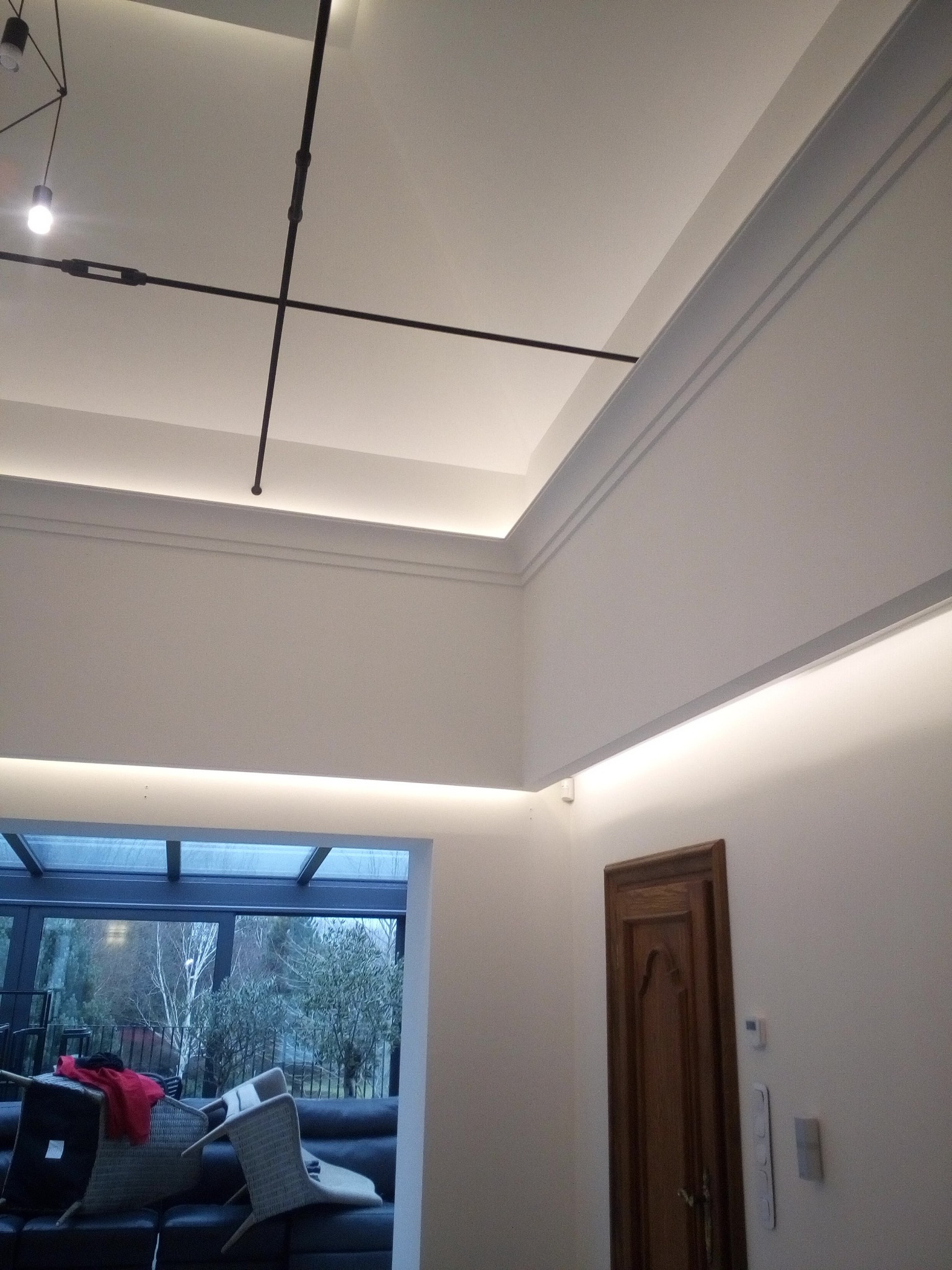 Nowoczesne wnętrze z podświetlanym sufitem, listwami dekoracyjnymi i minimalistycznym oświetleniem na tle szklanej ściany z widokiem na ogród