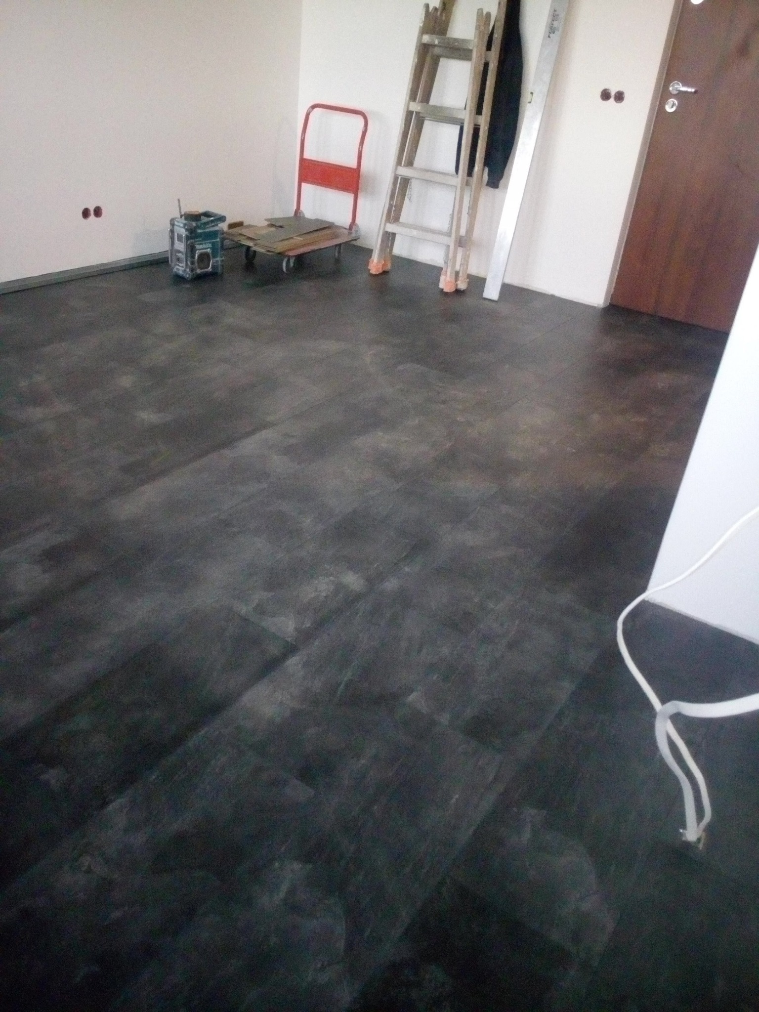 Ułożona podłoga z paneli w odcieniach szarości, widoczne narzędzia: drabina, wózek transportowy i radio budowlane, ściany przygotowane do dalszych prac wykończeniowych.