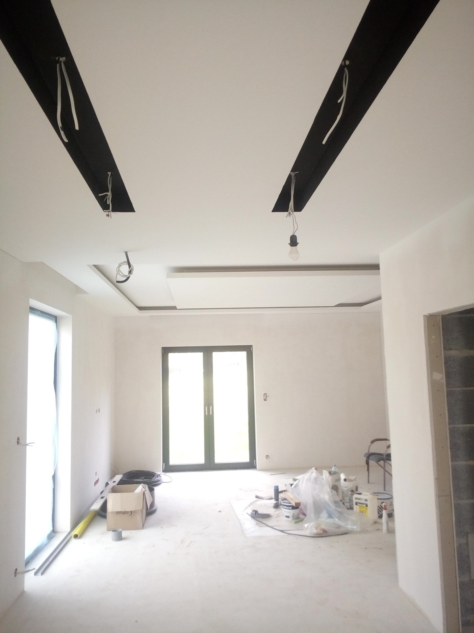 Wnętrze w trakcie wykańczania, widoczne czarne elementy dekoracyjne na suficie z wystającymi przewodami, ściany przygotowane do malowania, podłoga zabezpieczona, widoczne narzędzia i materiały...