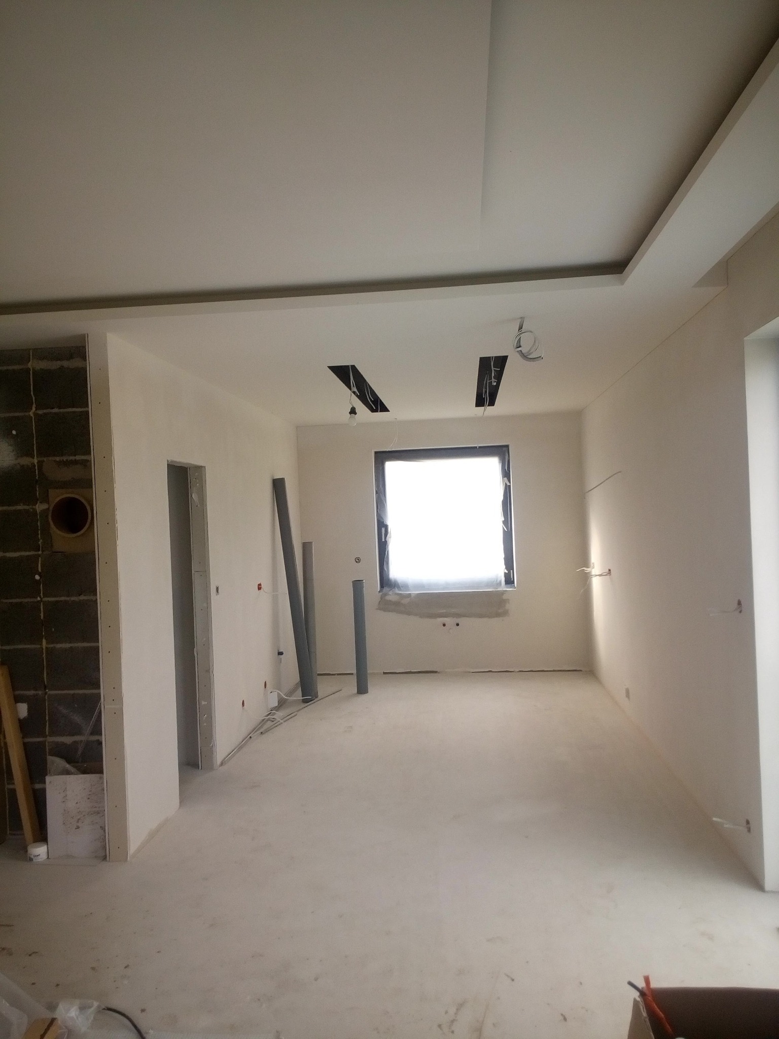 Wnętrze w trakcie prac wykończeniowych: surowy tynk na ścianach, widoczne instalacje elektryczne, okno zabezpieczone folią, przygotowane otwory w suficie pod oświetlenie.