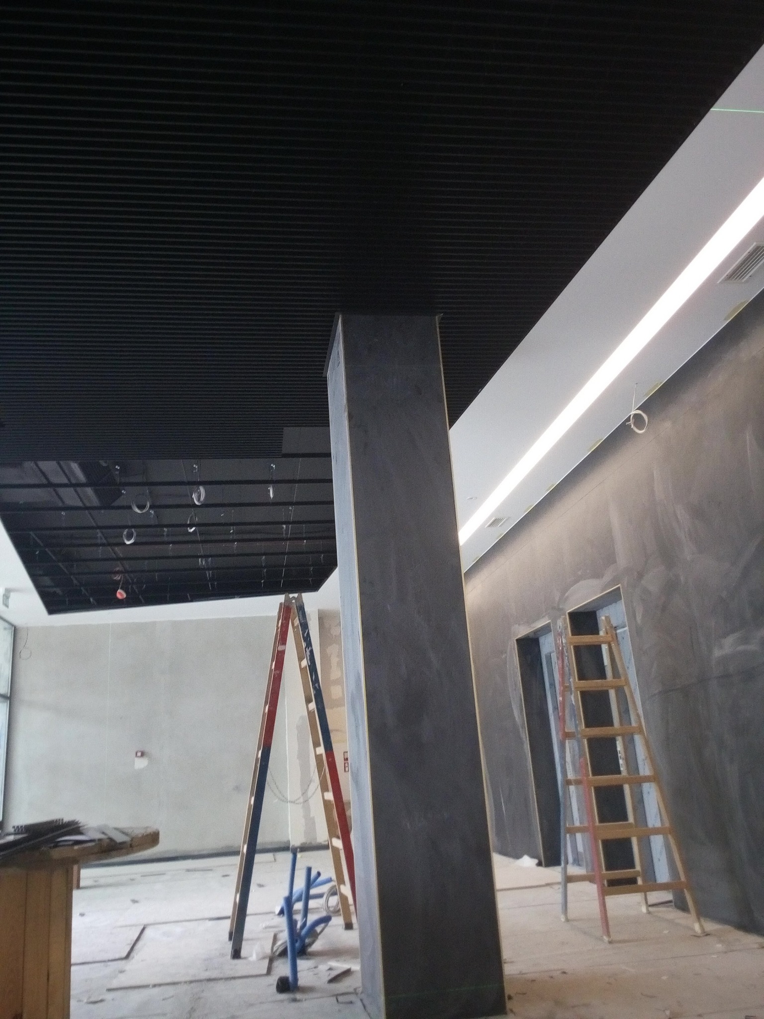 Wnętrze biura w trakcie prac wykończeniowych: widoczny sufit rastrowy, kolumna wykończona w szarym dekoracyjnym tynku, drabiny i narzędzia na podłodze.