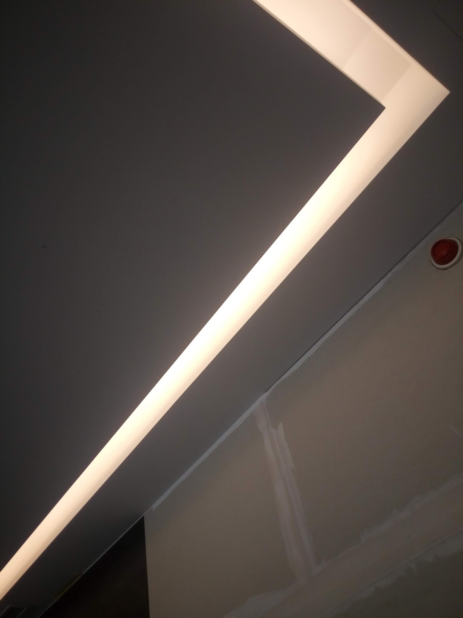 Kąt ujęcia sufitu z zamontowanym, liniowym oświetleniem LED w białej obudowie, widoczny fragment ściany z niedokończonymi szpachlowaniami i czujnik dymu.